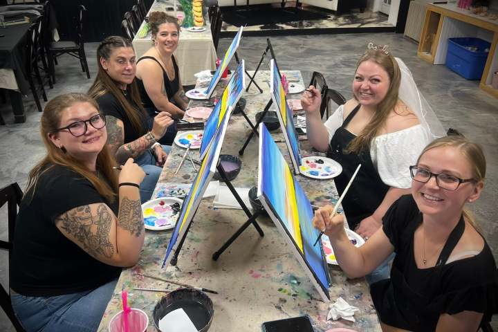 Five women painting at an art class, smiling at the camera, with easels and paint palettes.
