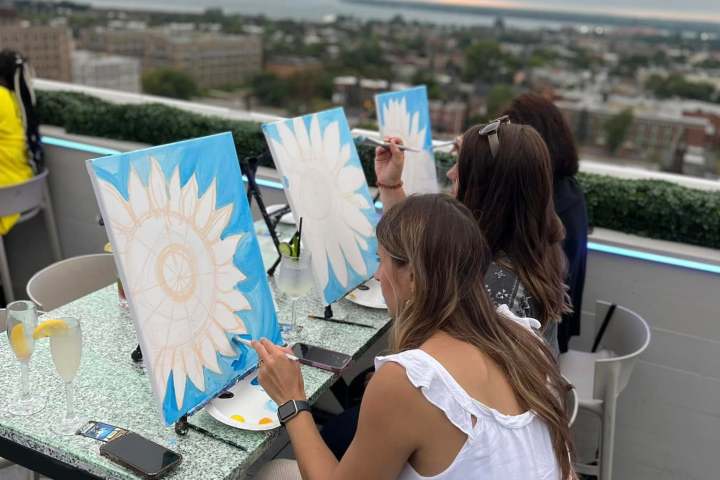 Three women painting sunflowers at an outdoor rooftop event with city view.