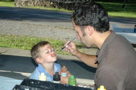 Man paints child's face outdoors at a table with art supplies.