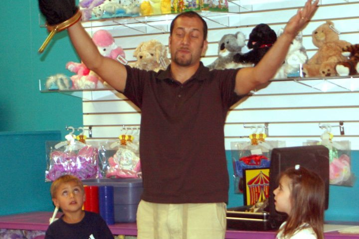 Man with puppet and children in toy store with stuffed animals on shelves.