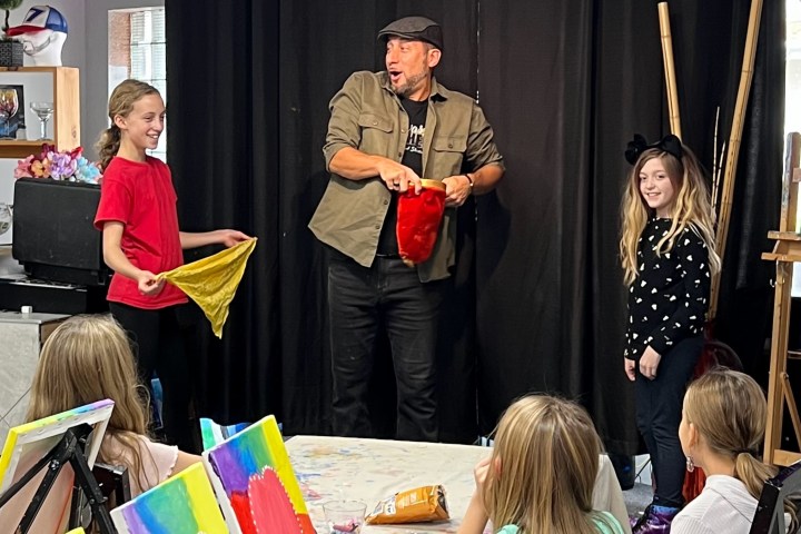 Man performing magic trick with two girls holding colorful bags on stage.
