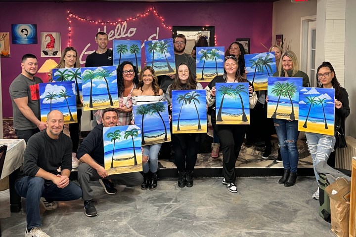 Group holding paintings of a beach scene with palm trees in a studio.