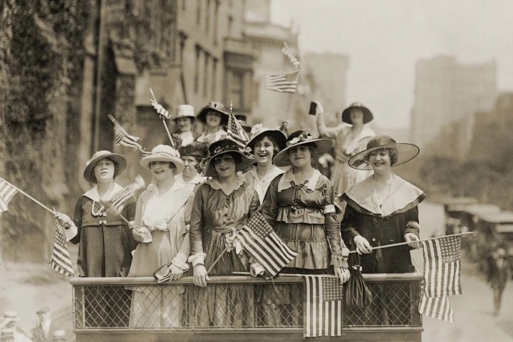 Group of women in vintage attire holding American flags on a vehicle in a city street.
