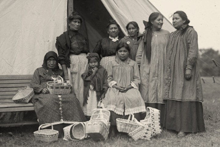 Seven women and a child in traditional clothing with woven baskets outside a tent.