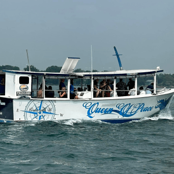 White boat named 'Queen of Peace' with passengers, sailing on a lake with trees in the background.